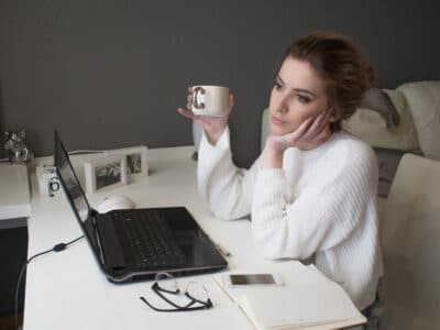 A woman looks out the window at her desk. She looks tired and her open laptop is on the desk in front of her.