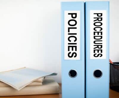 Two binders labeled "policies" and "procedures" are sitting on a desk next to a stack of papers.